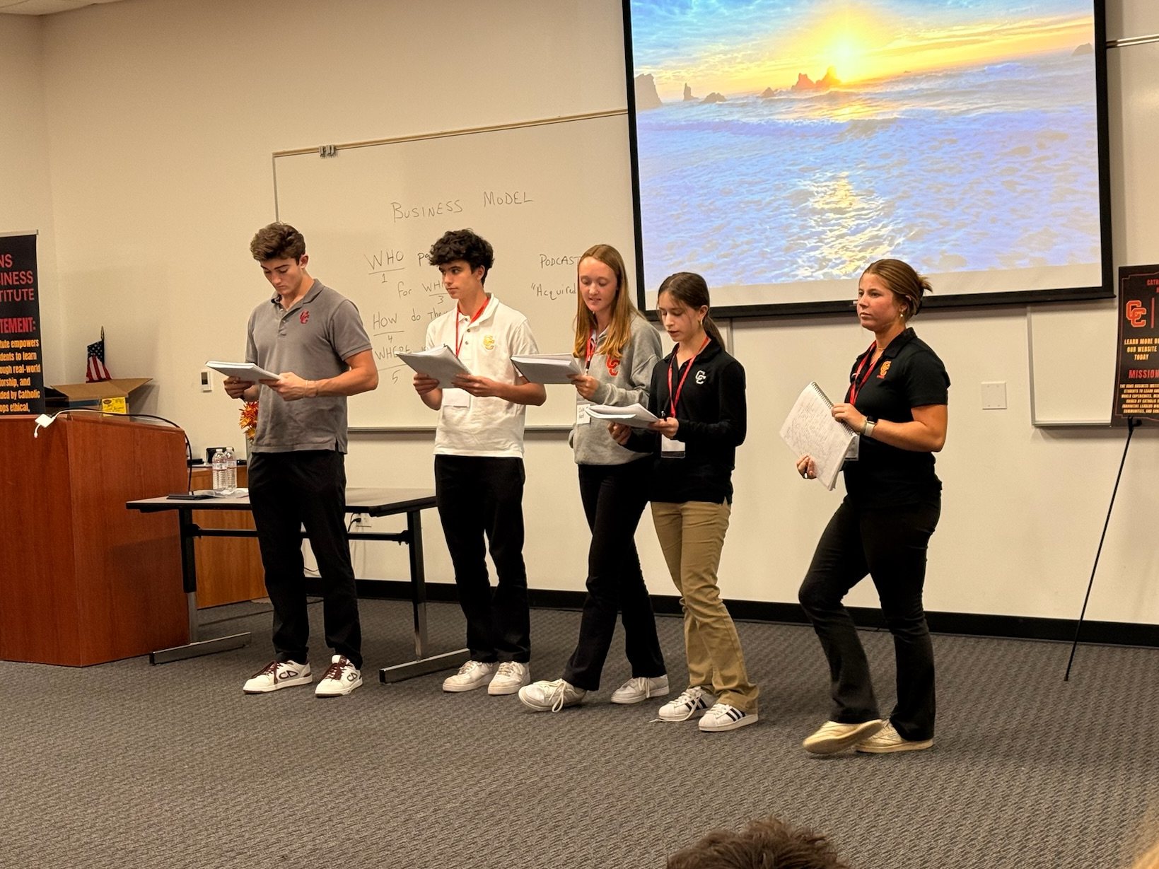 Five students presenting at the front of the room, a sunset slide on the screen and a business model sketched on the whiteboard.