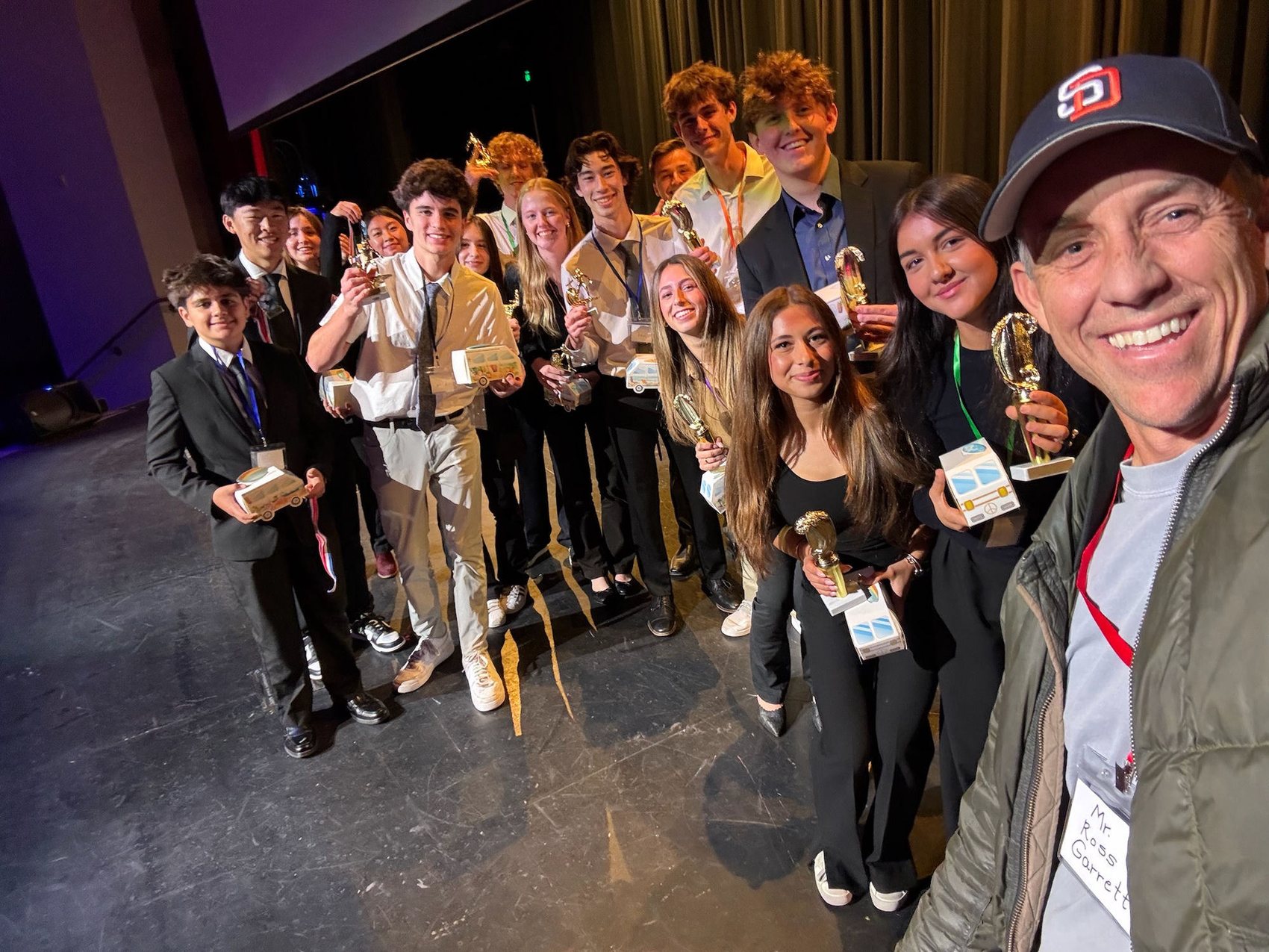 Surfline CEO Ross Garrett takes a selfie with the winning case competition students holding their trophies on stage.