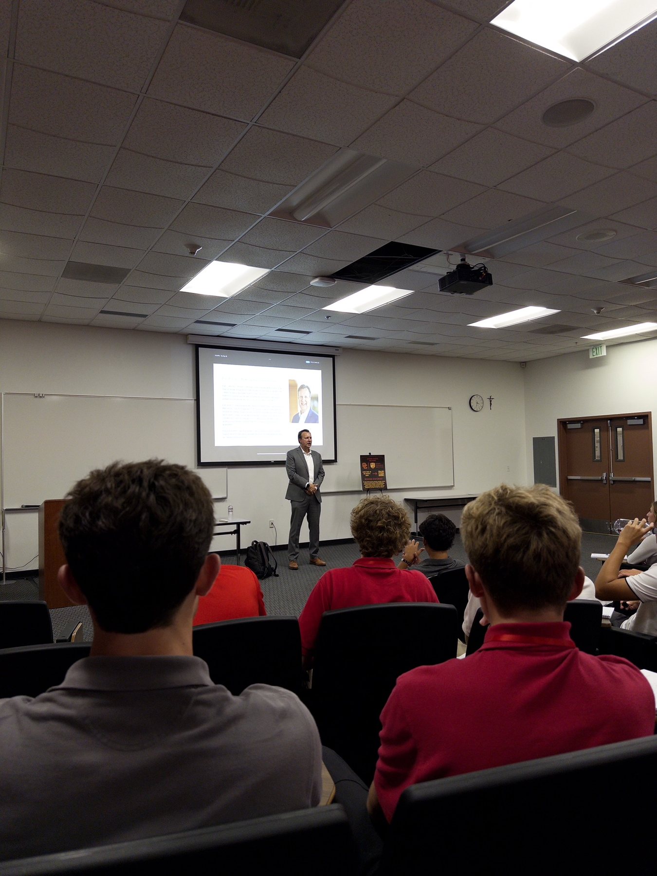 An industry speaker presenting to DBI students in red polos, a crucifix on the classroom wall.
