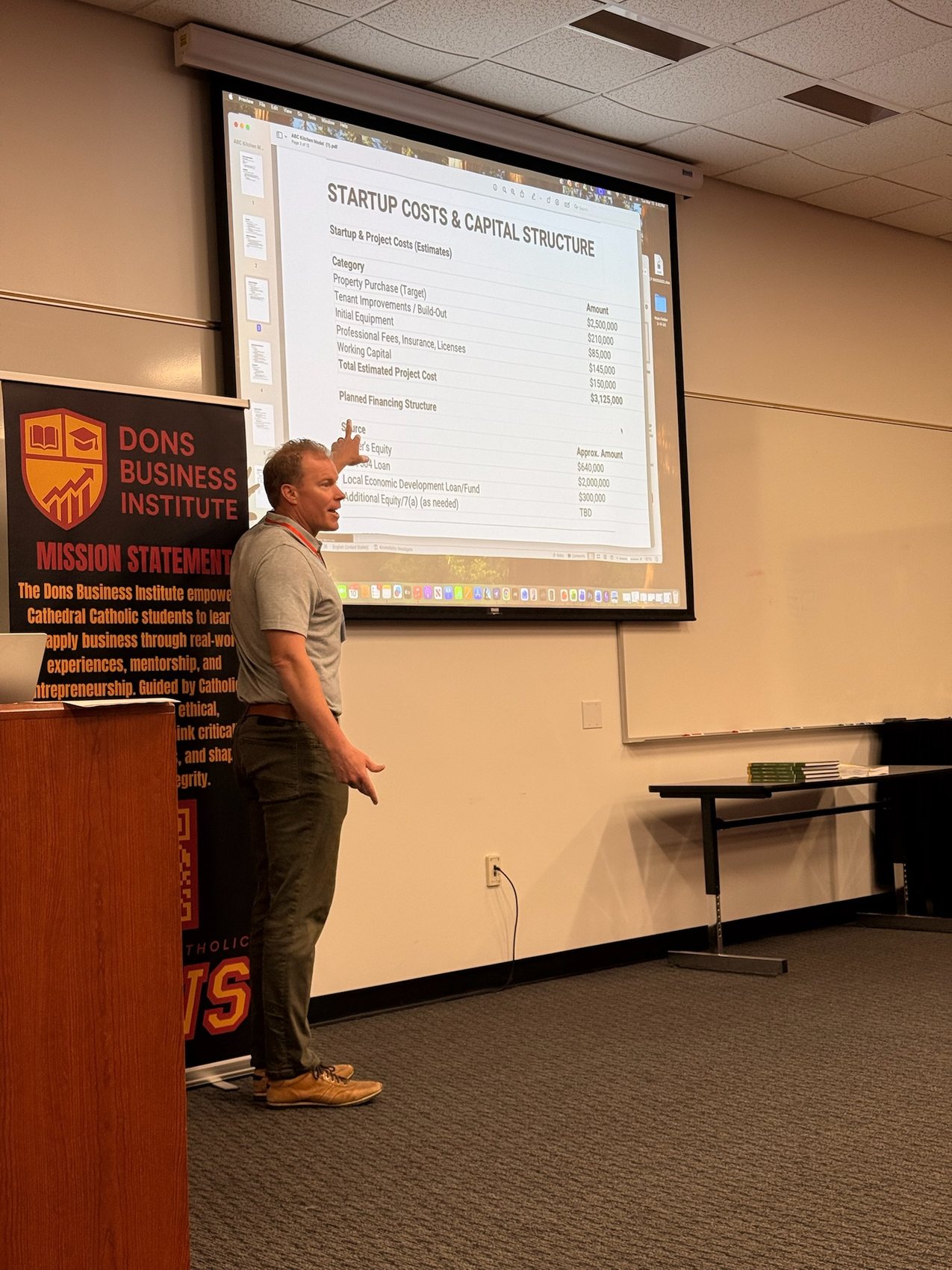 Adam Seery pointing to a startup costs and capital structure slide beside the DBI mission banner.